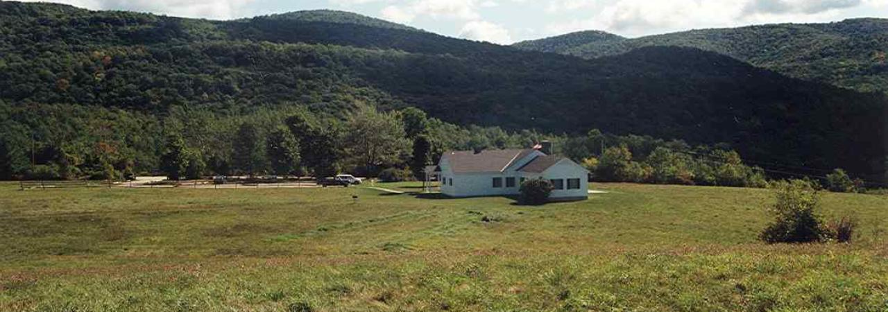 Photograph of a grassy battlefield with a white house in the mid-ground and a mountain range in the background.