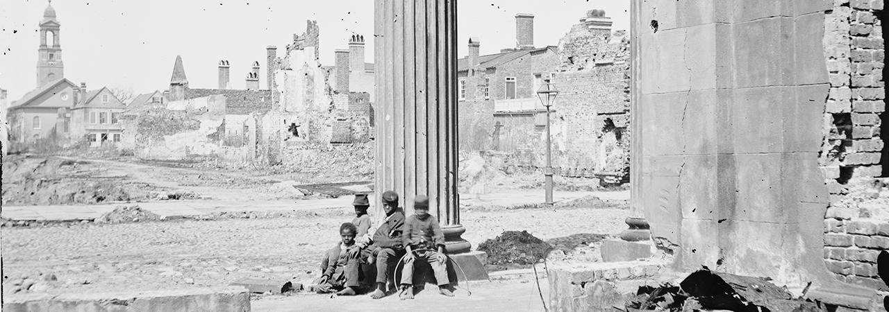 This is a black and white image of four young boys sitting barefoot and dressed in ragged clothing  on the rubble of a battlefield. 