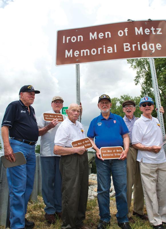 A group of eight veterans stands with a sign that reads Iron Men of Metz Memorial Bridge