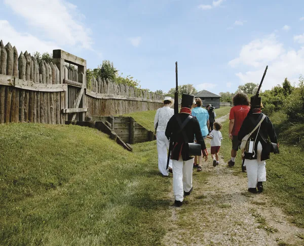 A group of tourists and two War of 1812 reenactors walk along a stone path next two a wooden fence.